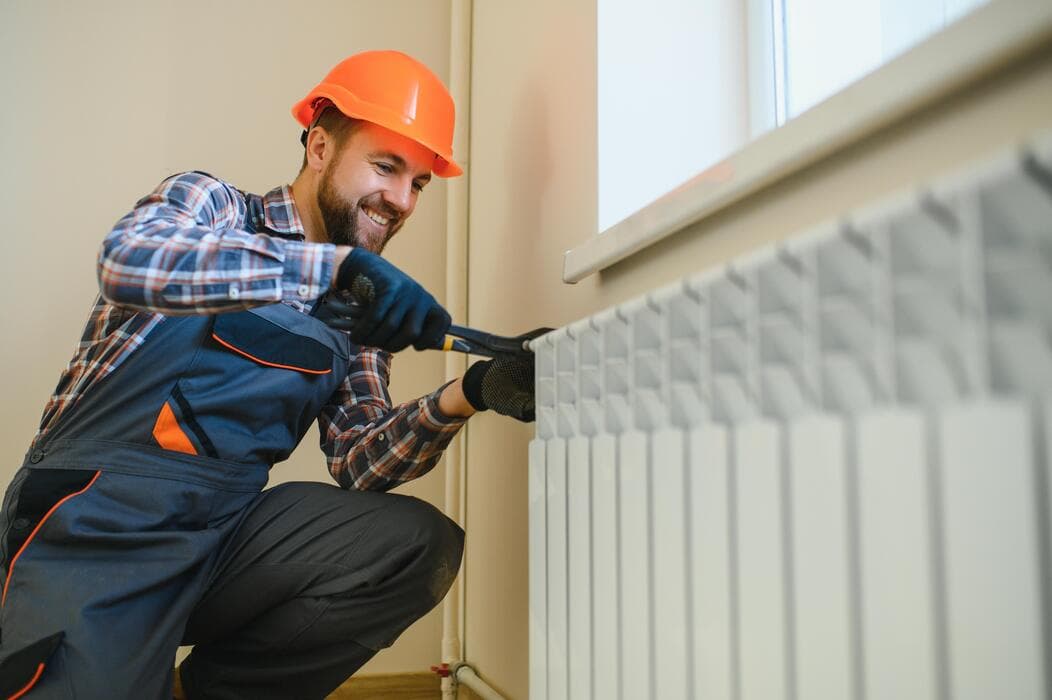 Installateur met helm die een radiator onder een raam aansluit met een sleutel
