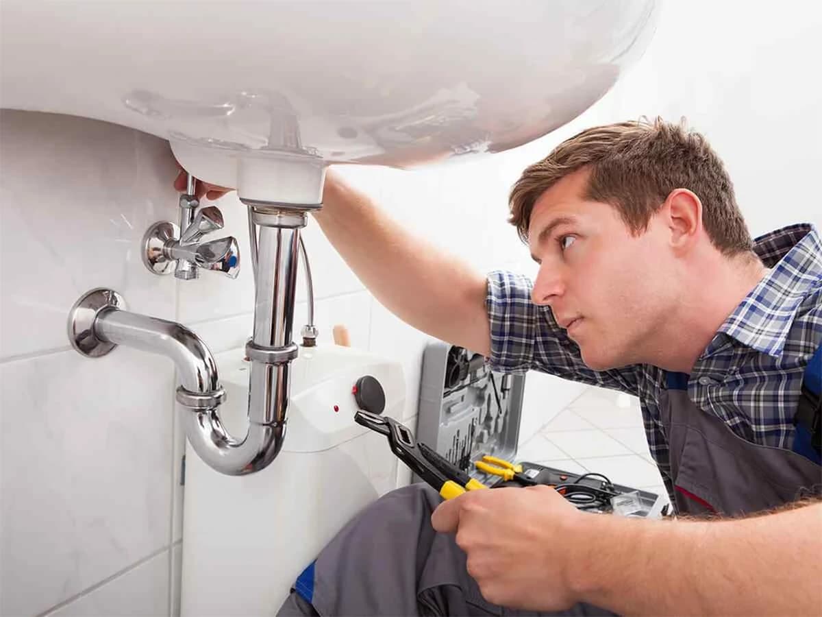 Loodgieter met een tang onder een wastafel, bij de sifon en een kleine boiler