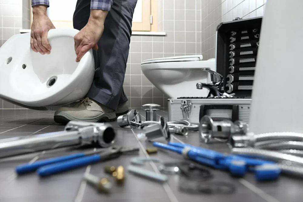 Man die een witte wastafel draagt in een betegelde badkamer, met een toilet, een open gereedschapskoffer en losse buizen, kranen en tangen op de vloer