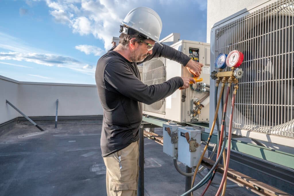 Homme en casque blanc qui travaille sur l'unité extérieure d'une pompe à chaleur sur un toit plat, avec des manomètres sur les raccords